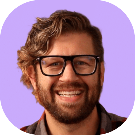 A smiling man with wavy brown hair, a beard, and black glasses is shown against a light purple background. He is wearing a plaid shirt, embodying the relaxed spirit of Highline 2025.