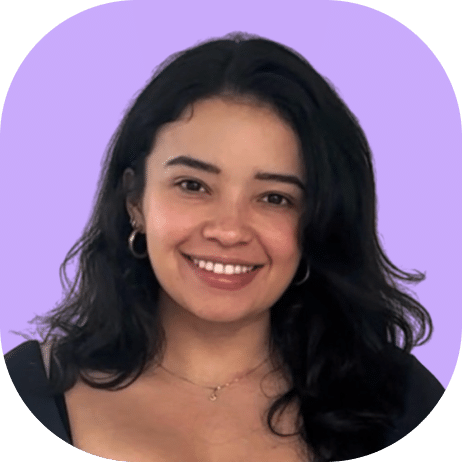 A woman with long, wavy dark hair smiles at the camera. She is wearing gold hoop earrings, a necklace, and a dark top. The background is a solid light purple color with rounded corners.